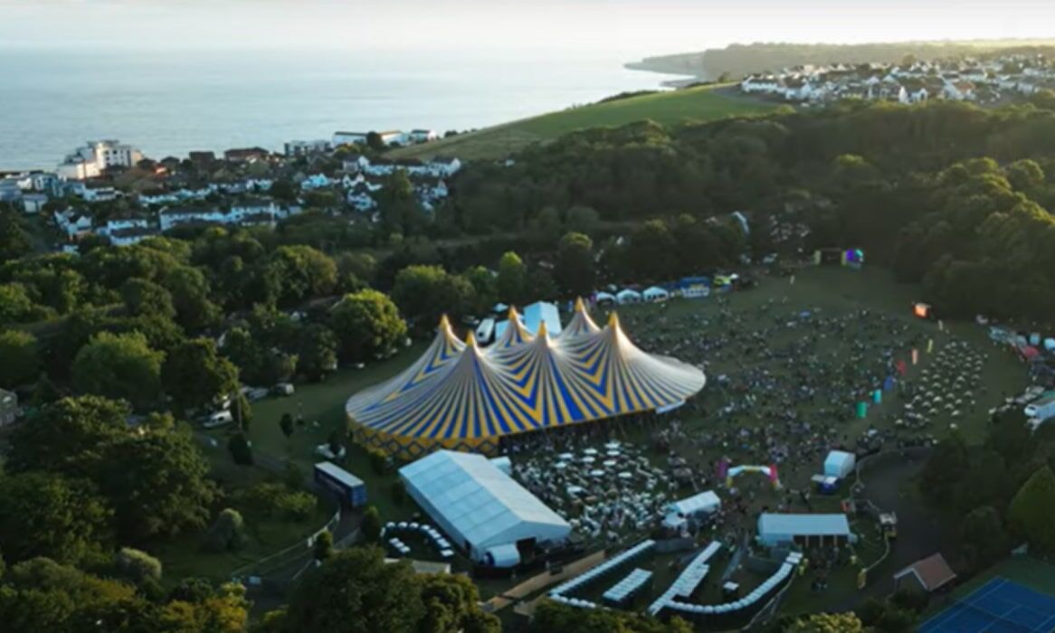 Aerial View Over GlastonBarry's colorful festival main yellow and blue Tent, surrounding forestry, and the coast of Barry.